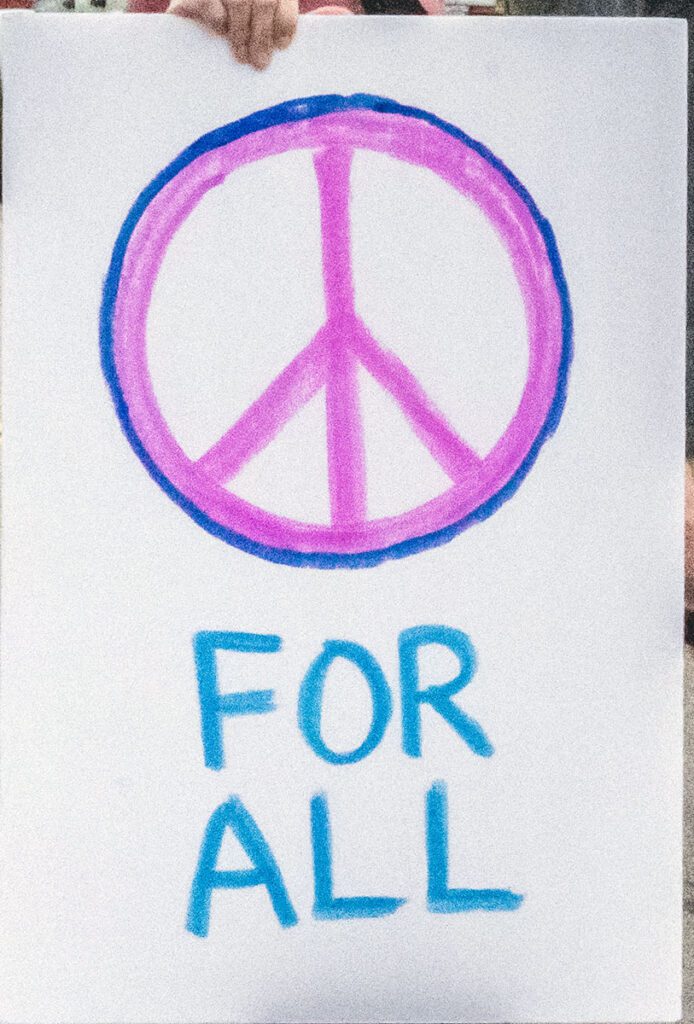 A hand holds a sign with a colorful peace symbol in pink and blue above the words "FOR ALL" written in blue letters.