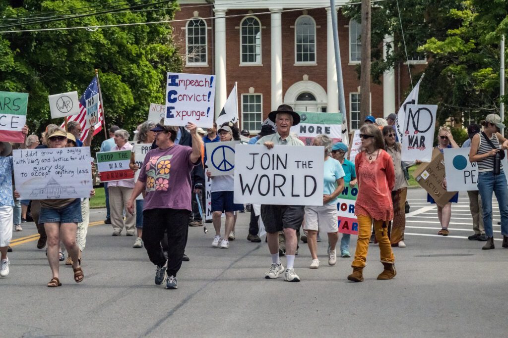 A group of people participating in a peaceful protest march down a street, carrying signs with anti-war messages. One sign reads "JOIN THE WORLD," and another reads "Impeach! Convict! Expunge!" in front of a building with columns and trees in the background.