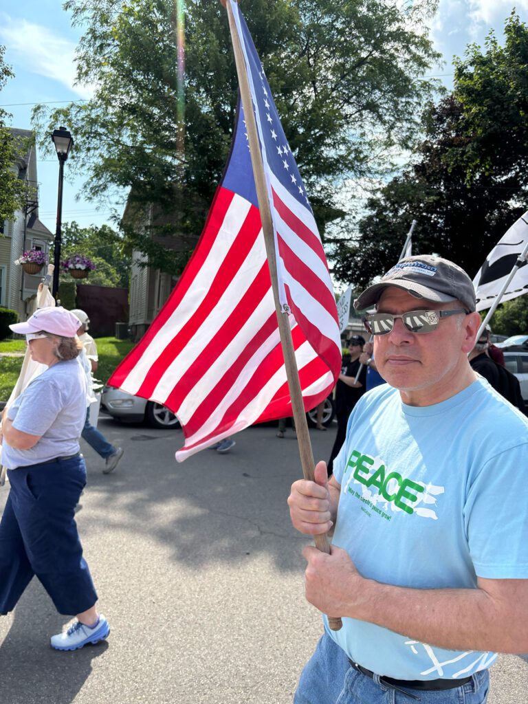 A person wearing sunglasses and a light blue "PEACE" T-shirt holds an American flag during a sunny outdoor event. Other participants walk nearby, and trees and houses are visible in the background.