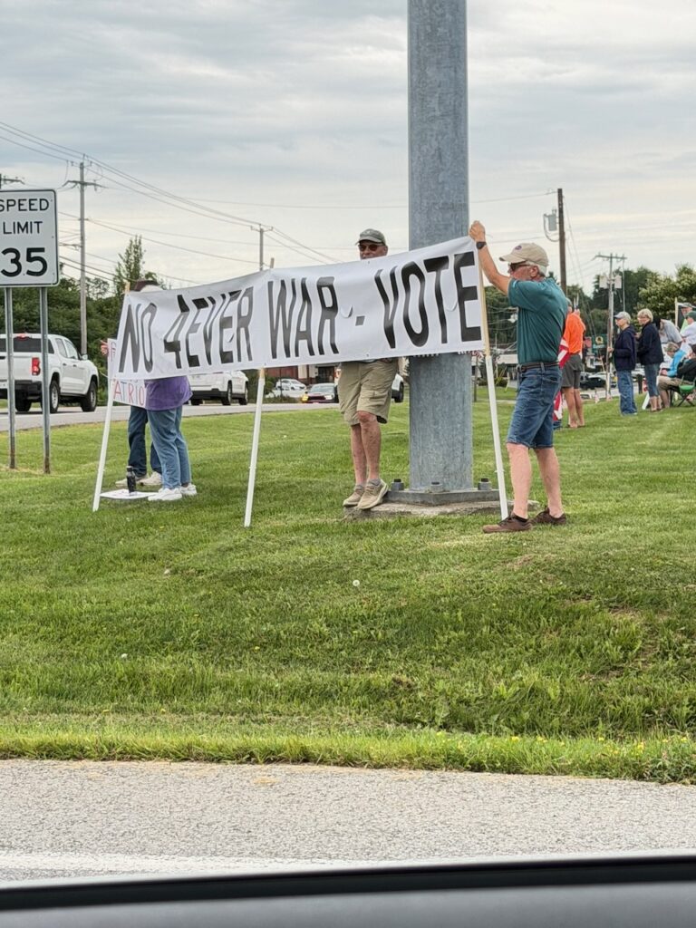 Protesters holding 'No Forever War - Vote' banner on roadside
