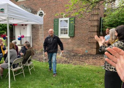 A person walks on a lawn towards a small gathering seated under a canopy, while others stand and clap near a brick building with green shutters. The event appears celebratory, with decorations in the background.