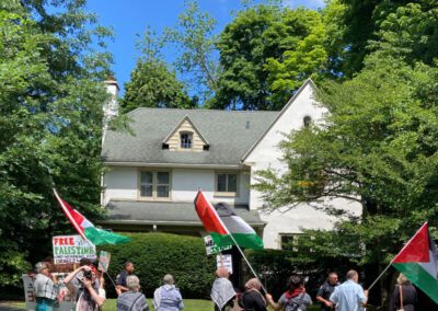 A group of people holding Palestinian flags and banners gather outside a white house surrounded by trees on a sunny day.