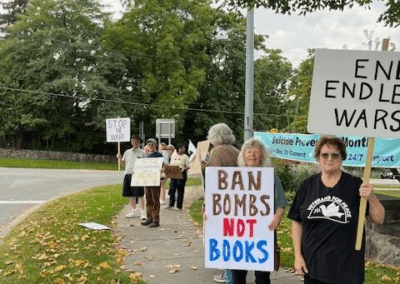 A group of people standing on a sidewalk holding protest signs, including one that reads "BAN BOMBS NOT BOOKS" and another saying "END ENDLESS WARS." The scene is set outdoors with trees and a street in the background.