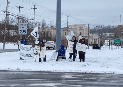 A group of people holding signs advocating for peace stand by a snowy roadside, with messages like "Buy Peaceful Toys" and "Peace on Earth."