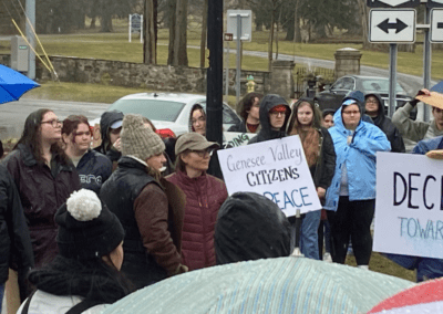 A group of people standing in an outdoor setting, holding signs with messages like "Genesee Valley Citizens for Peace"