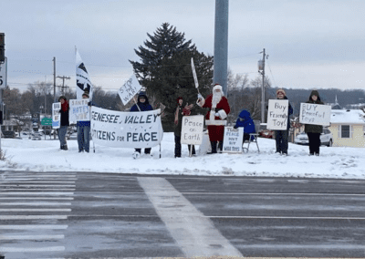 A group of people stand on a snowy street corner holding signs advocating for peace, with one person dressed as Santa Claus. The signs include messages such as "Stop War," "Peace on Earth," and "Genesee Valley Citizens for Peace."