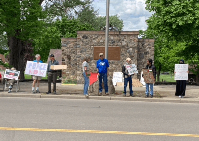 A group of people holding protest signs stand in a line near a stone memorial and trees. Signs include messages like "Peace Now," "Ceasefire Now," and "No Apartheid, Free Palestine."