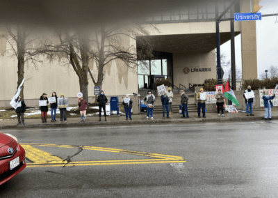 A group of protesters holding signs stand on the sidewalk in front of an L3 Harris building. Some signs mention Israel, and a Palestinian flag is visible. The scene is overcast, with some patches of snow on the ground.