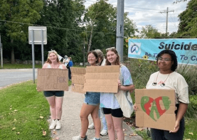 Four young people standing on a sidewalk holding cardboard signs with various messages, participating in an outdoor event. A banner related to a suicide prevention campaign is visible in the background.