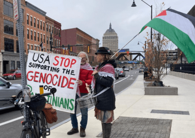 Two people stand on a city street holding a large sign that reads, "USA Stop Supporting the Genocide of Palestinians". One person holds a Palestinian flag, and a bicycle is parked nearby with buildings and traffic visible in the background.