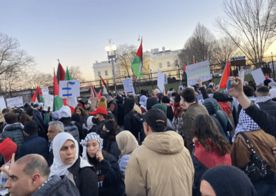 A large group of people protesting outside a white building, many holding flags and signs, some wearing keffiyehs and headscarves.