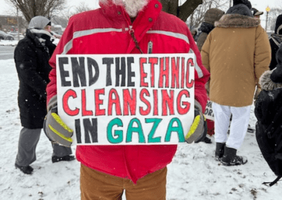 A person in a red jacket holding a sign that reads "END THE ETHNIC CLEANSING IN GAZA" stands in a snowy outdoor setting with others in the background.
