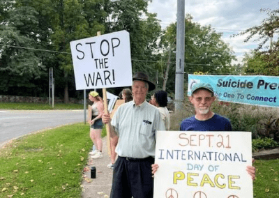 Two men standing on a sidewalk holding signs advocating peace; one reads "STOP THE WAR!" and the other says "SEPT. 21 INTERNATIONAL DAY OF PEACE" with peace symbols. A green lawn and trees are in the background.