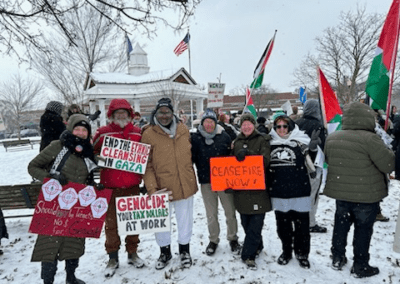 A group of people standing in a snowy area, holding signs protesting for a ceasefire and against actions in Gaza. The signs include messages like 'End the Ethnic Cleansing in Gaza' and 'Ceasefire Now!' with some Palestinian flags visible in the background. They are dressed warmly for winter weather.