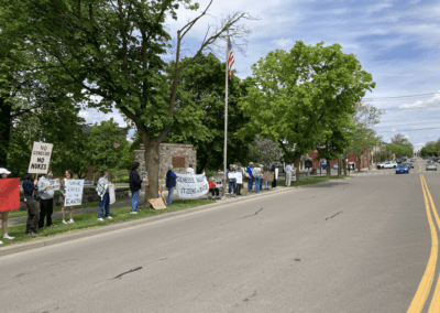 A group of people standing on the sidewalk holding signs with messages advocating against genocide, nuclear weapons, and war in a peaceful protest near a street lined with trees and a flagpole.