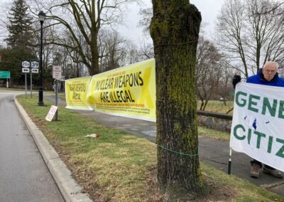 A protest scene with banners stating "Nuclear Weapons Are Illegal" and other messages, displayed in a park area with trees and grass. A man is holding part of the banner to the right.