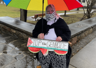 A person sits on a foldable chair holding a sign reading "Stop the Genocide" in front of them, under a multicolored umbrella. They are wearing a patterned scarf over their head and a black and white outfit. The setting is a rainy park or outdoor area with wet pavement and bare trees.