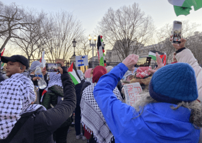 A group of people at a protest holding Palestinian flags and signs, some wearing keffiyehs, with phones raised to film the event; trees and a building are visible in the background.