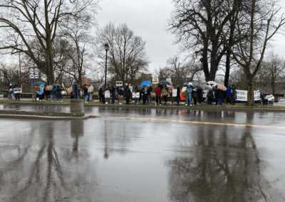 A group of people holding protest signs gather on a rainy street, with bare trees and wet pavement visible.
