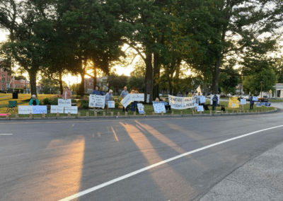 A group of people standing along a roadside holding protest signs advocating for peace and the abolition of nuclear weapons, with trees and buildings in the background.