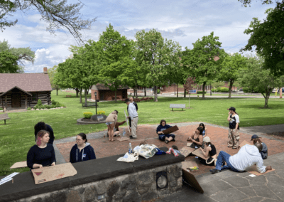 A group of people gathered in a park setting with green trees and a clear sky, some sitting on the ground and others standing, engaged in an outdoor activity with cardboard signs.