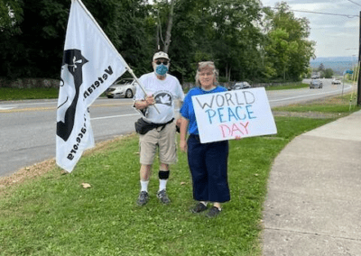 Two people stand on a grassy roadside, one holding a flag with a graphic design, and the other holding a sign that reads 'WORLD PEACE DAY.'