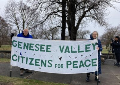 Two elderly people holding a banner that reads "Genesee Valley Citizens for Peace" in a park with bare trees; a person with a camera stands nearby.