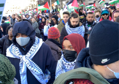 A crowded protest with people wearing scarves, hats, and masks, holding Palestinian flags in a city street.