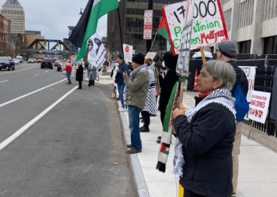A group of people stand on a city sidewalk holding signs and flags in a protest. One person holds a large flag with red, green, white, and black vertical stripes. Other participants hold signs, one of which reads "17,000+ Palestinian Children Martyred" in green and red letters. Buildings are visible in the background along the street.