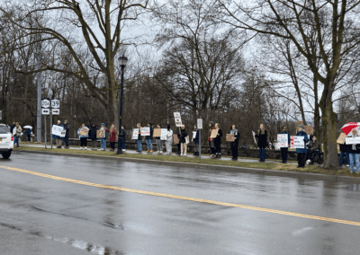 A group of people standing along a roadside holding protest signs, under overcast skies with damp road conditions, surrounded by leafless trees.