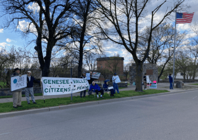 A peaceful protest with people holding banners and signs for peace, including one that reads 'Genesee Valley Citizens for Peace,' in an outdoor setting with trees and a flying American flag.