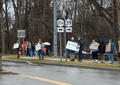 A group of people standing on a roadside in a wooded area, holding various protest signs including messages about decency and opposing certain actions.