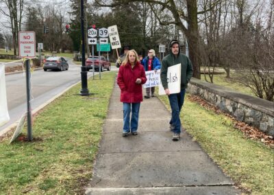 People walking on a sidewalk holding protest signs, with trees and a street in the background.