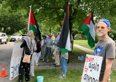 A group of people gathered outside, holding Palestinian flags and signs advocating for peace. One sign reads 'never again for anyone.' The scene includes trees, a sidewalk, and parked cars, with a peaceful protest atmosphere.