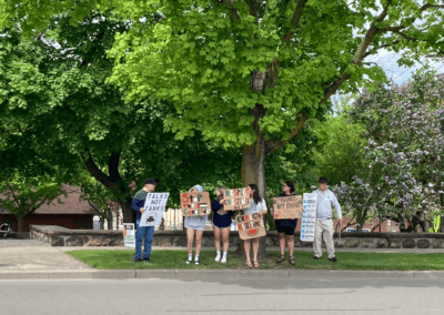 A group of people holding protest signs stand under a large, leafy tree. The signs include messages such as "Talks not tanks," "Stop the genocide," and "Respect existence or expect resistance." They are participating in a peaceful protest on a grassy area next to a road.