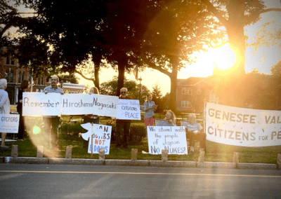 A group of people standing and sitting outside holding signs advocating for peace and remembrance of Hiroshima and Nagasaki; the sunlight shines through trees in the background.