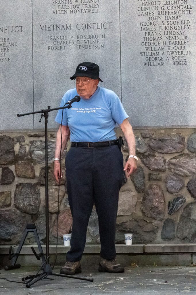 An elderly man wearing a blue T-shirt with "Genesee Valley Citizens for Peace" and a website address, stands in front of a memorial wall with engraved names and "Vietnam Conflict" inscribed on it. He is speaking into a microphone on a stand and wearing a black hat with an emblem, surrounded by stonework. Two paper cups are visible on the stone ledge behind him.