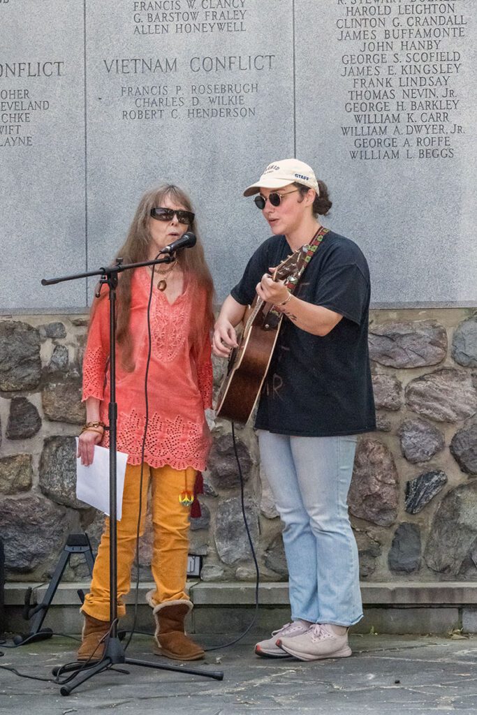 Two people performing in front of a memorial wall, with one singing into a microphone and the other playing a guitar.