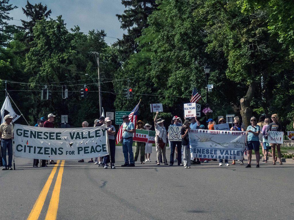 A group of protestors stand on a street holding banners and signs, including one reading 'Genesee Valley Citizens for Peace,' in a tree-lined area.