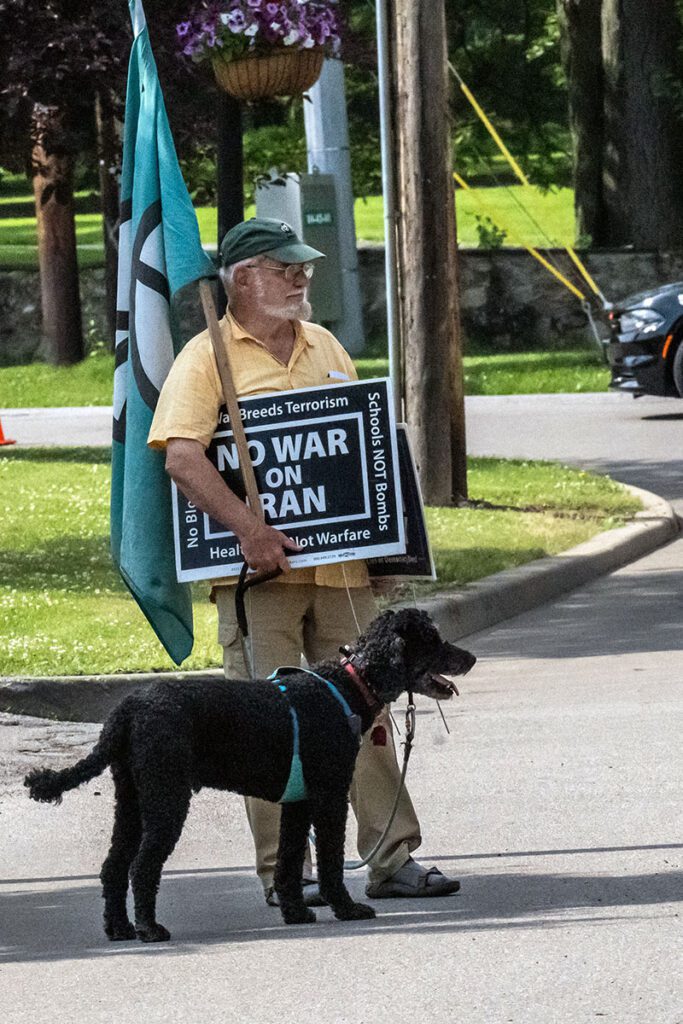 A man holding a protest sign and flag stands on a street with a black dog on a leash beside him. The sign reads 'No War on Iran' among other messages advocating against war and promoting peace.
