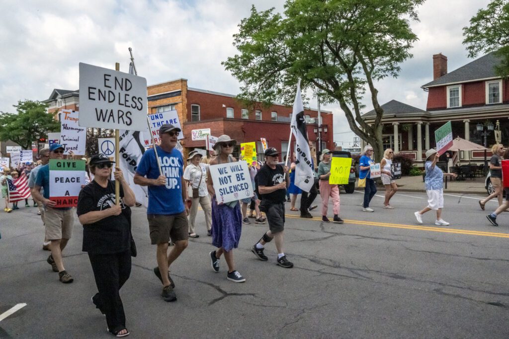 A group of protesters marching down a street holding signs with messages like "End Endless Wars," "Give Peace a Chance," and "We Will Not Be Silenced." The participants are diverse in age and the setting includes brick buildings and trees.