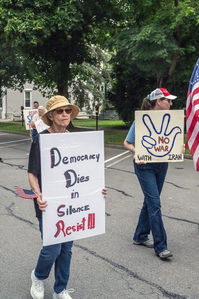 A woman wearing a sun hat holds a sign reading "Democracy Dies in Silence, Resist!!!" during a protest. Nearby, another person carries a sign with a peace symbol hand gesture, reading "No War with Iran." An American flag is partially visible.