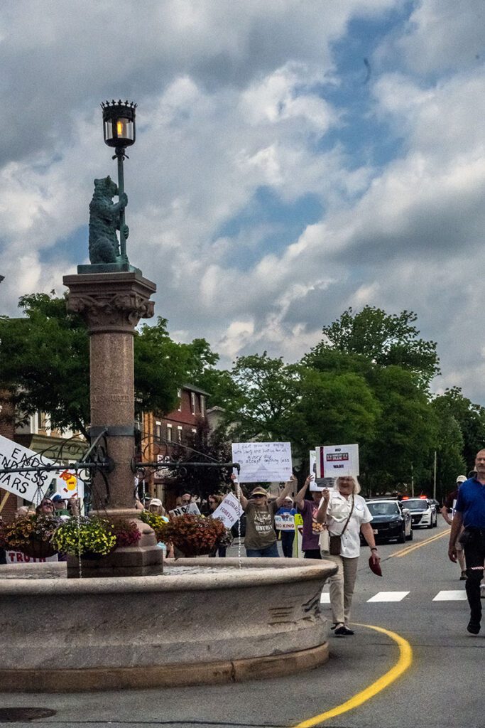 A group of protesters holding signs walk near a fountain with a statue and lamppost. The cloudy sky and green trees form the background.