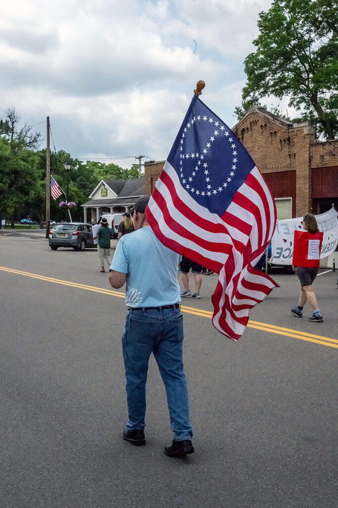 A person in a light blue shirt and jeans holds a flag featuring a peace symbol within a circle of stars, reminiscent of a Betsy Ross flag, walking in a parade.