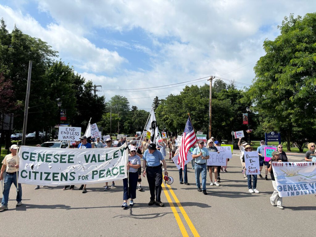 A group of protesters marching down a street holding various signs and banners, including one that reads "Genesee Valley Citizens for Peace" and another with an American flag. The sky is partly cloudy, and trees line the sides of the road.